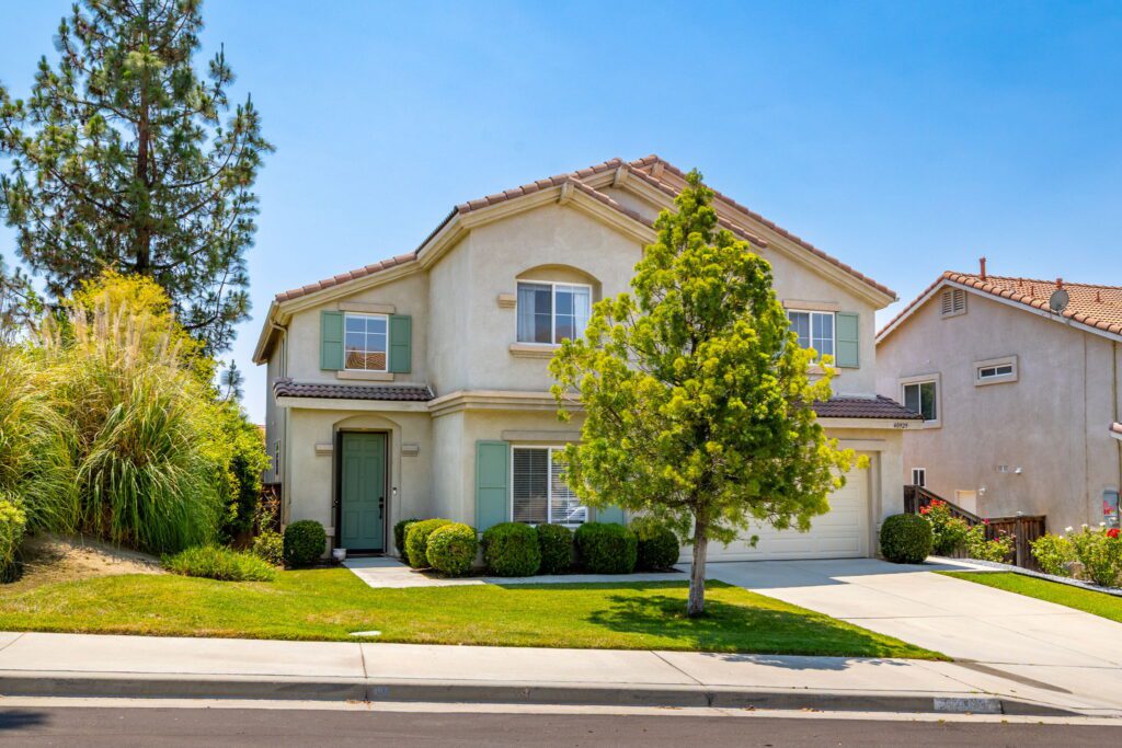 Two-story residential home with green shutters and a double garage, surrounded by well-kept landscaping under a clear blue sky.