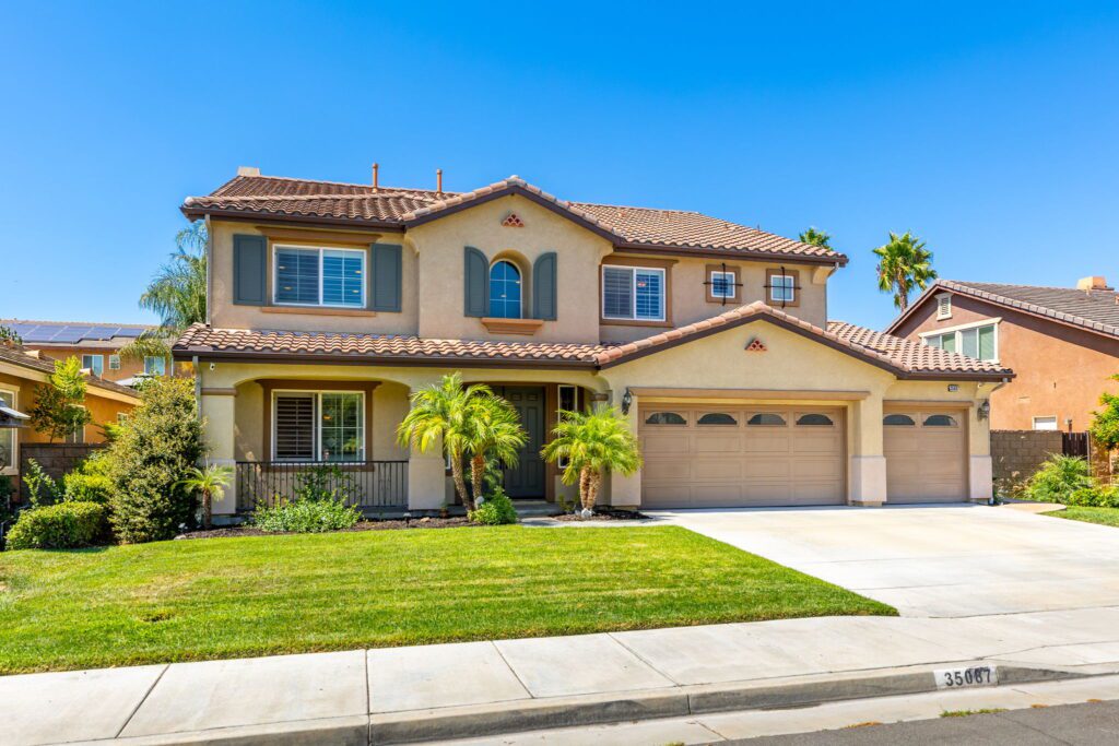 Two-story suburban house with a tile roof, green shutters, and palm trees in the yard. The garage has two doors, showcasing a well-maintained facade.