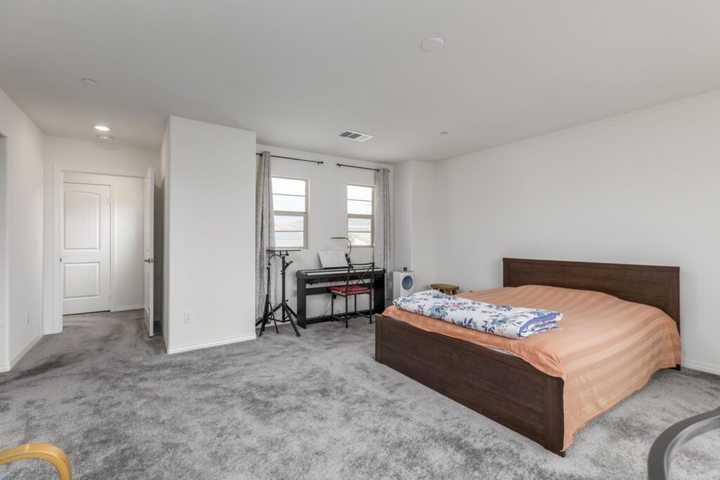Bright, spacious bedroom with a bed, patterned blankets, and a keyboard setup in the corner. Windows allow natural light, enhancing the welcoming atmosphere.