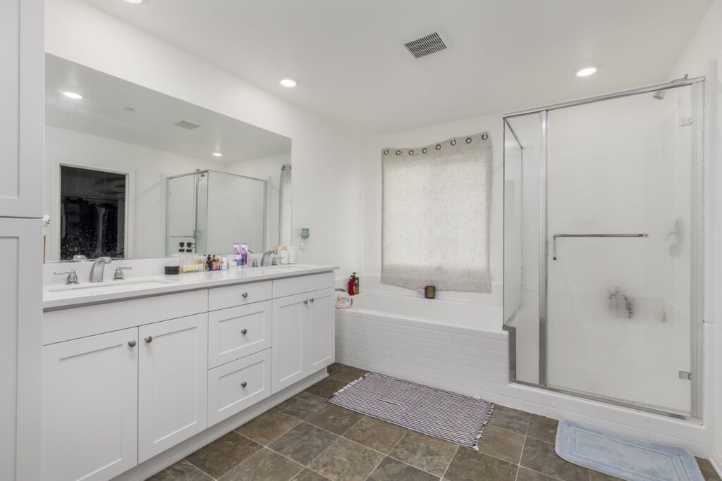 Modern bathroom featuring a double vanity, a glass-enclosed shower, and a bathtub. Natural light filters through a curtained window.