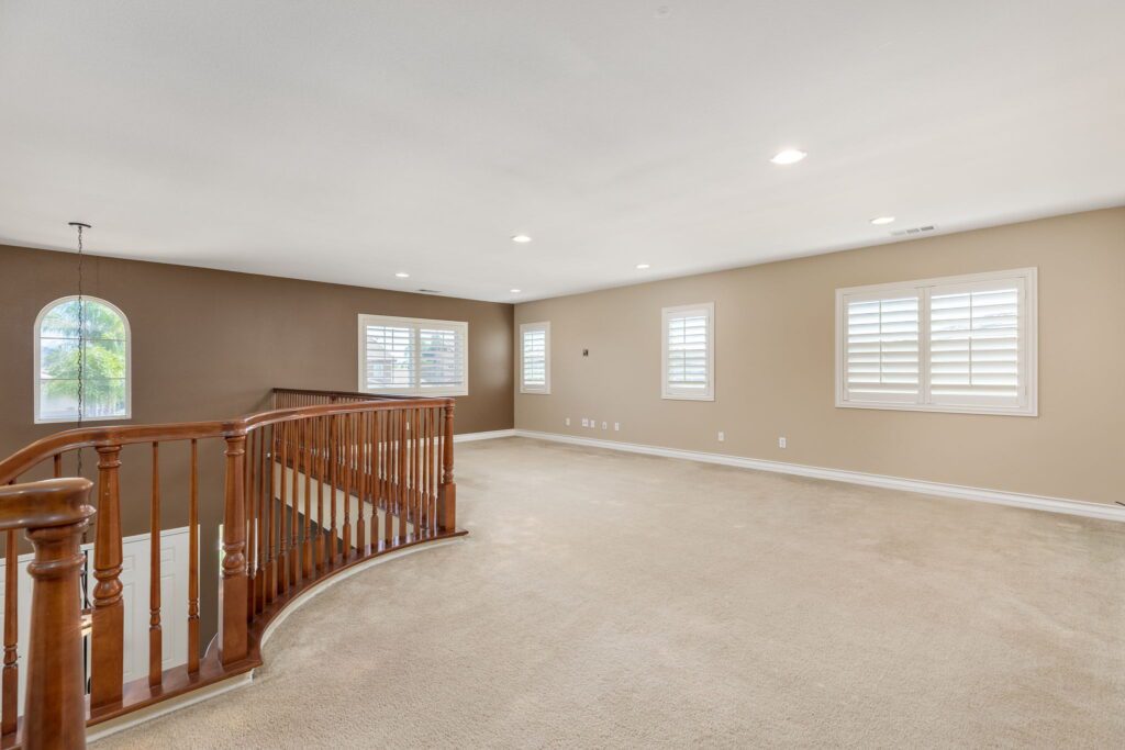 Spacious, well-lit upstairs living area features plush carpeting, a wooden rail, and multiple windows with shutters, creating a cozy atmosphere.