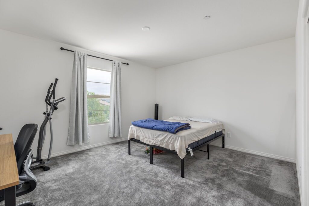 Bright, minimalist bedroom featuring a bed with unmade linens, a blue blanket, an exercise machine, and a desk. Natural light floods the space.