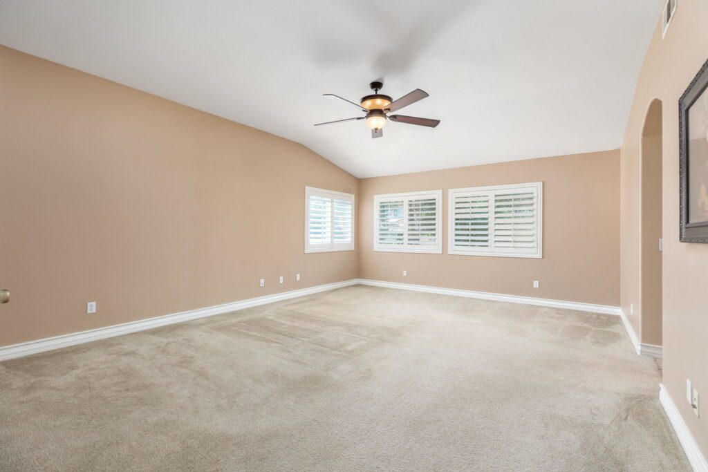 Spacious living room with tan walls and plush carpet. Features a ceiling fan and window shutters, providing a bright and airy atmosphere.
