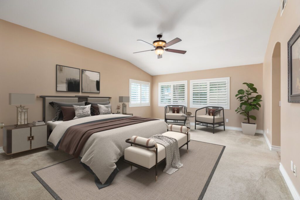 Stylish, modern bedroom featuring a queen bed with neutral-toned bedding, armchairs, and a ceiling fan. Natural light filters through plantation shutters.
