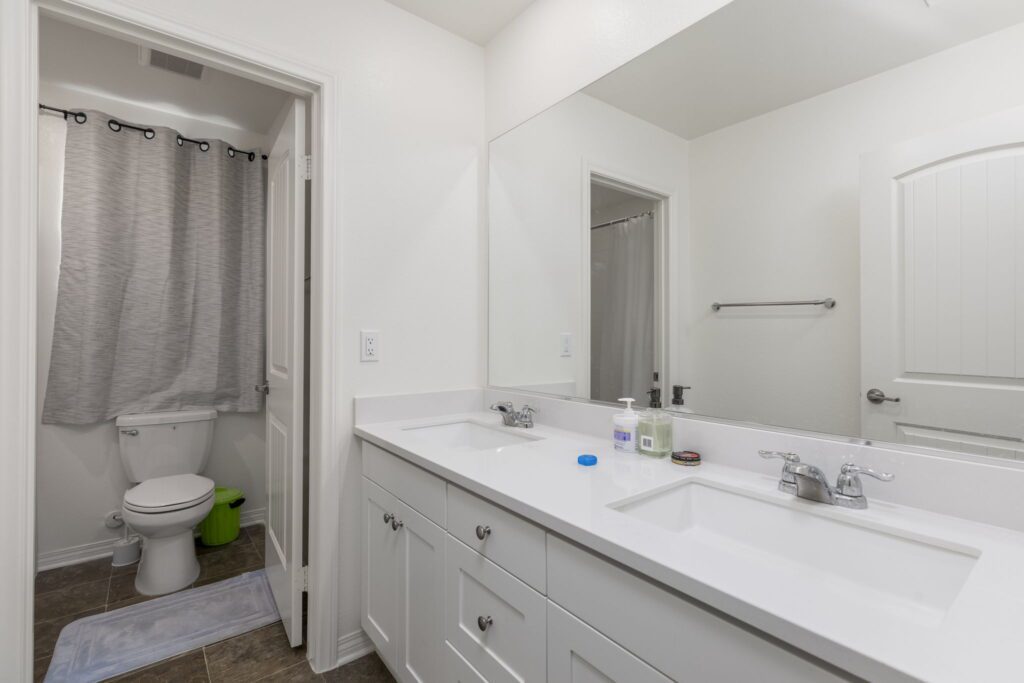 Modern bathroom featuring a double vanity with sinks, a large mirror, and a toilet hidden behind a gray curtain. Clean and well-lit design.