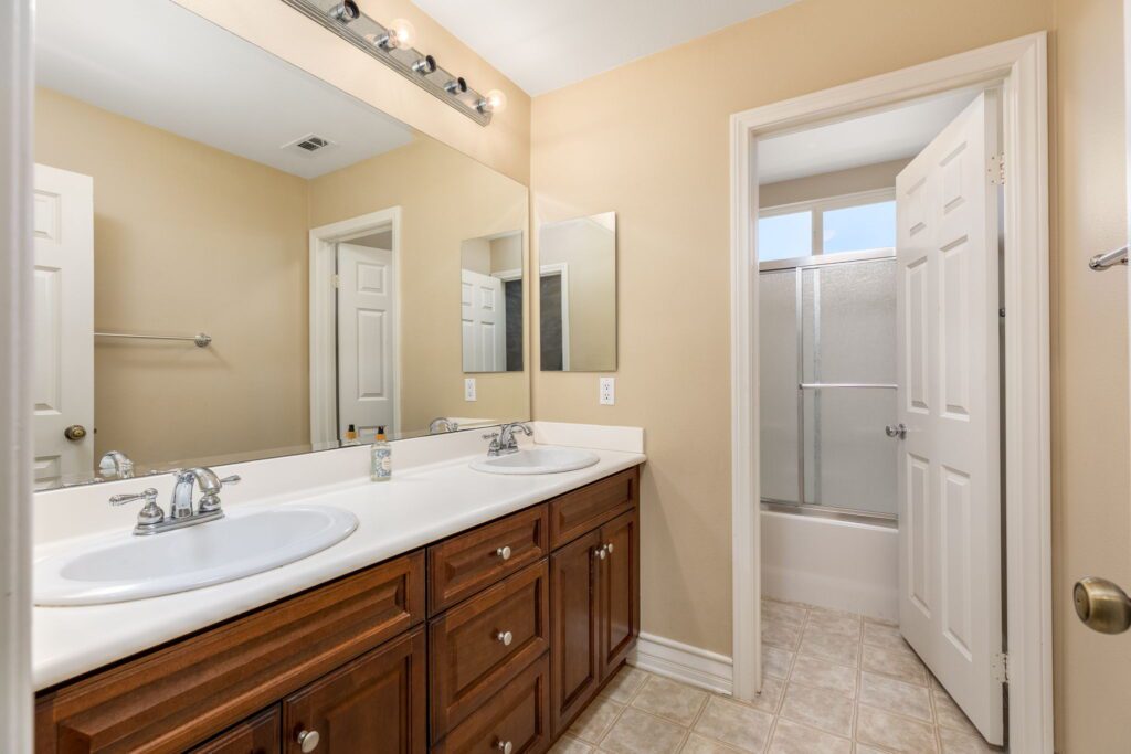 Bright, modern bathroom featuring a double sink vanity with wooden cabinetry, two mirrors, and a shower area visible through an open door.