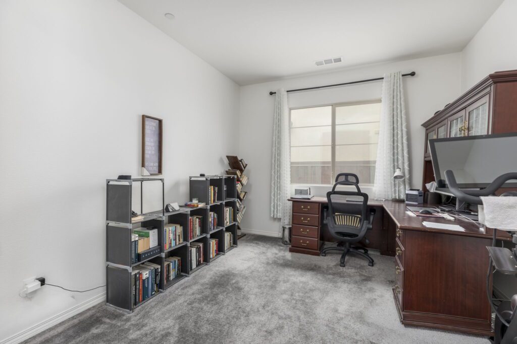 Modern home office featuring a spacious desk with a computer, an ergonomic chair, and a bookshelf filled with books. Natural light filters through a large window.