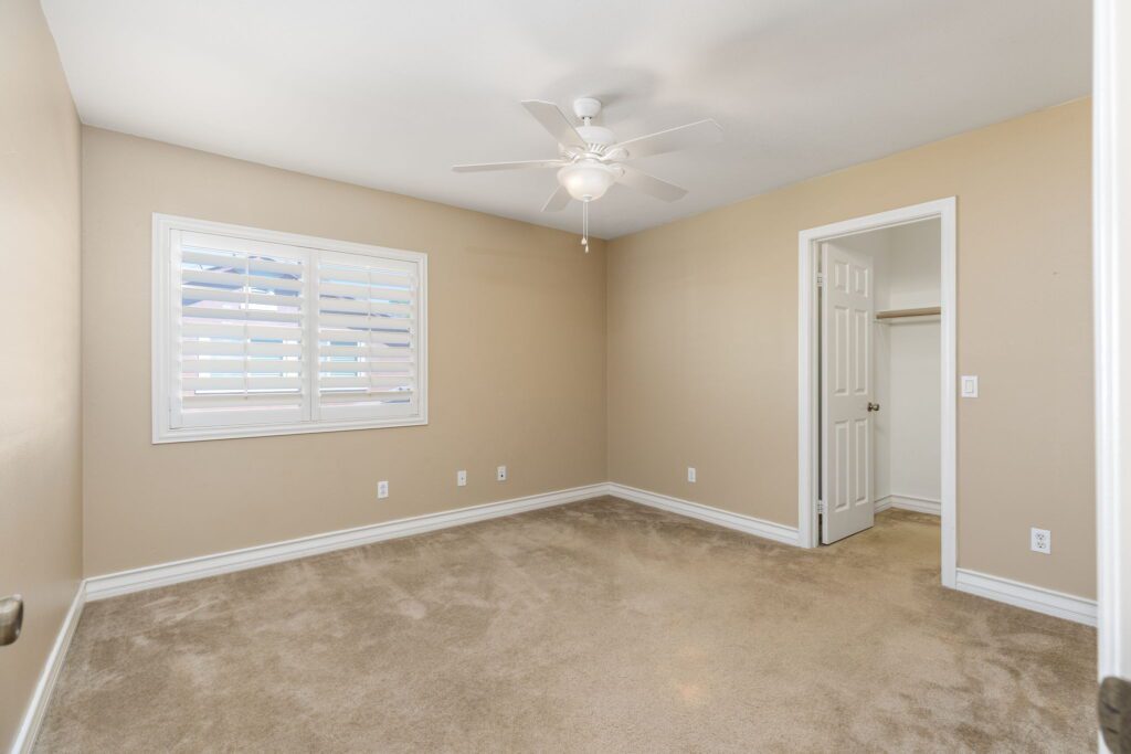 Spacious, neutral-toned bedroom featuring carpeted flooring, a ceiling fan, a window with shutters, and a closet. Ideal for home listings.