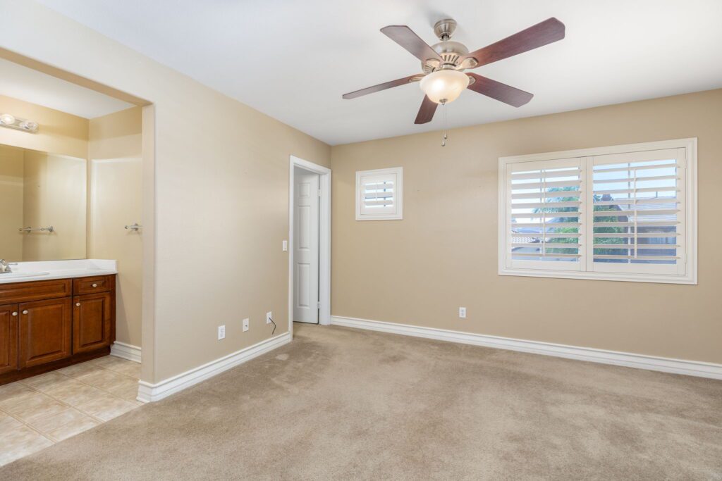 Spacious bedroom featuring a ceiling fan, plush carpet, light-colored walls, and a window with shutters, creating a bright, inviting atmosphere.