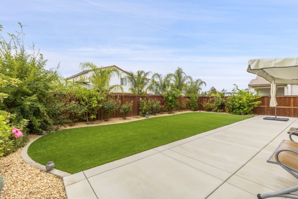 Lush green lawn bordered by colorful shrubs and a gravel pathway, with a patio area featuring a light umbrella and chairs, under a clear sky.