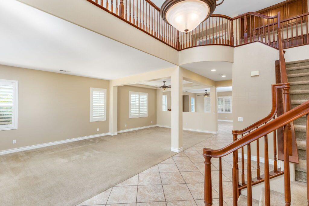 Spacious living area with beige walls, plush carpet, and tile flooring. Features a stairway with wooden banister and large windows letting in natural light.