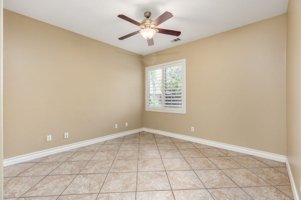 Empty room featuring beige walls, tiled flooring, and a ceiling fan. A window with shutters allows natural light, enhancing the spacious feel.