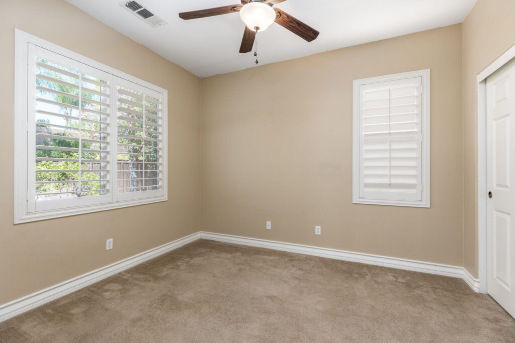 Empty room featuring beige walls, a light ceiling fan, and two windows with shutters. The carpeted floor enhances the cozy atmosphere.