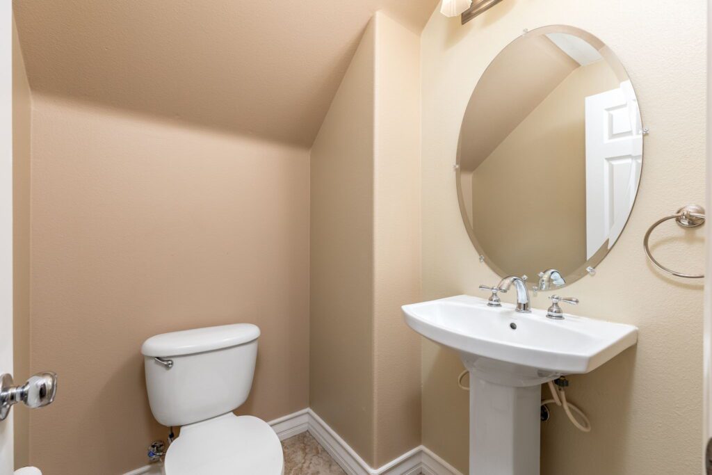 Small, well-lit bathroom featuring a white toilet and pedestal sink with chrome fixtures. An oval mirror reflects the space, emphasizing its compact design.