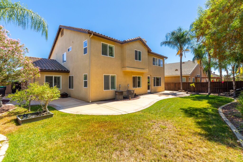 Two-story house with beige stucco exterior, surrounded by a well-maintained yard featuring palm trees and flowering shrubs. Ideal for outdoor living.