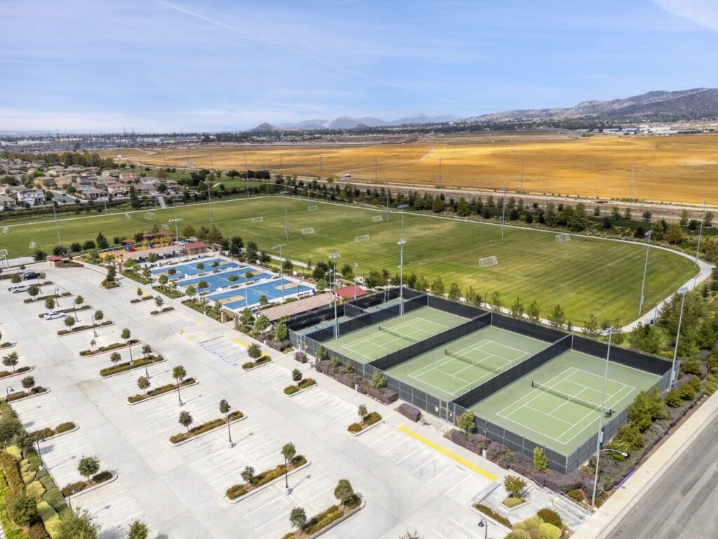 Aerial view of a sports complex featuring multiple tennis courts, basketball courts, and a large soccer field, surrounded by parking and residential areas.