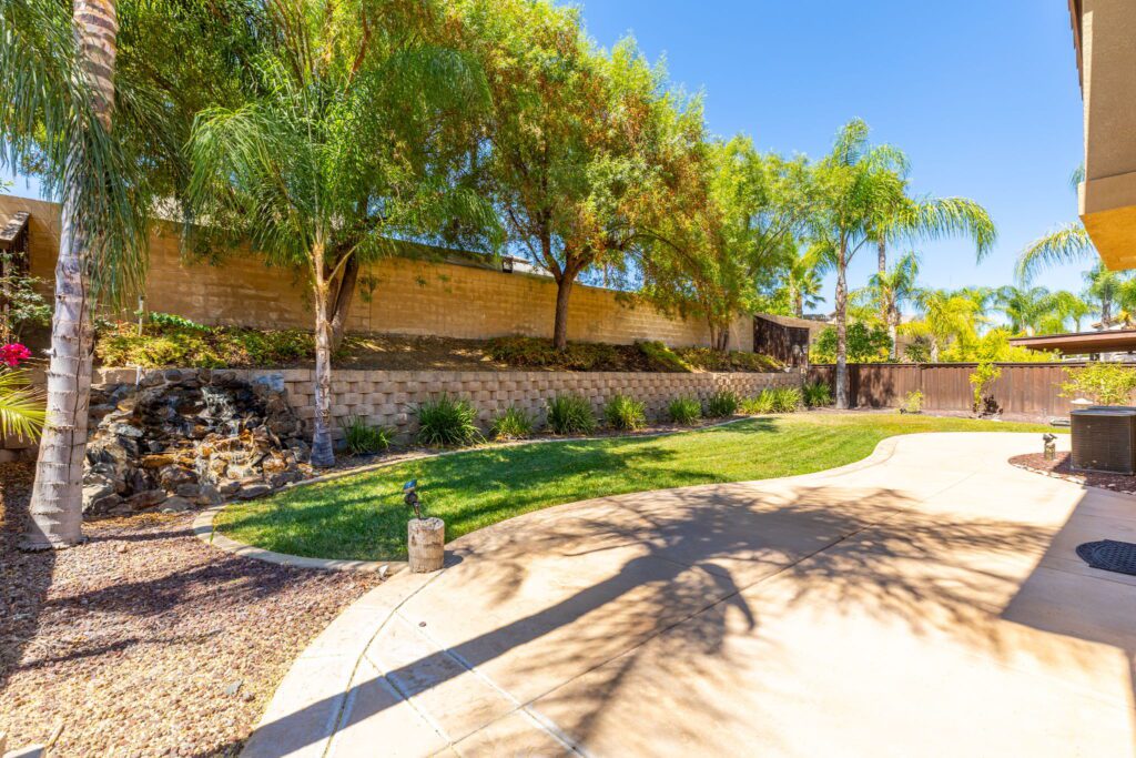 Sunlit backyard featuring a manicured lawn, palm trees, and a small stone waterfall. The curved pathway adds charm to the serene landscape.