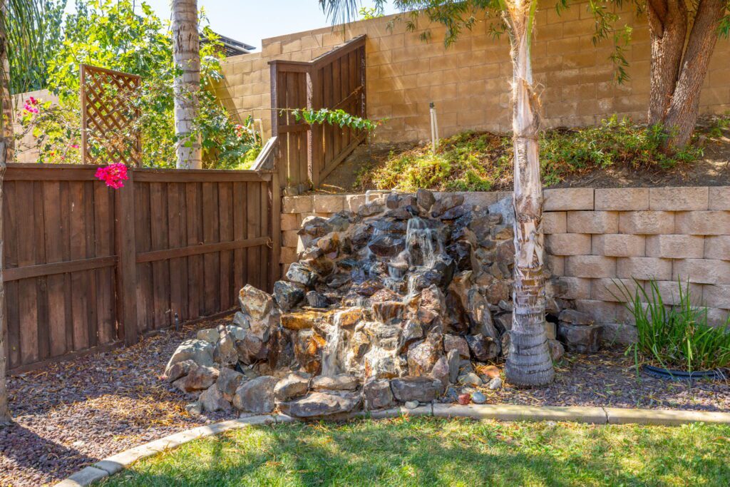 Tranquil backyard scene featuring a rock waterfall surrounded by greenery and a wooden fence, enhancing the outdoor aesthetic.