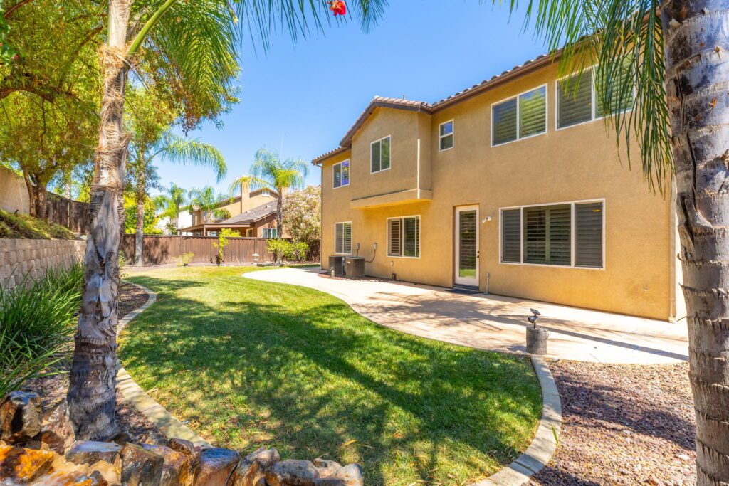 Backyard view of a two-story home featuring a patio, manicured lawn, and palm trees under clear blue skies, ideal for outdoor gatherings.
