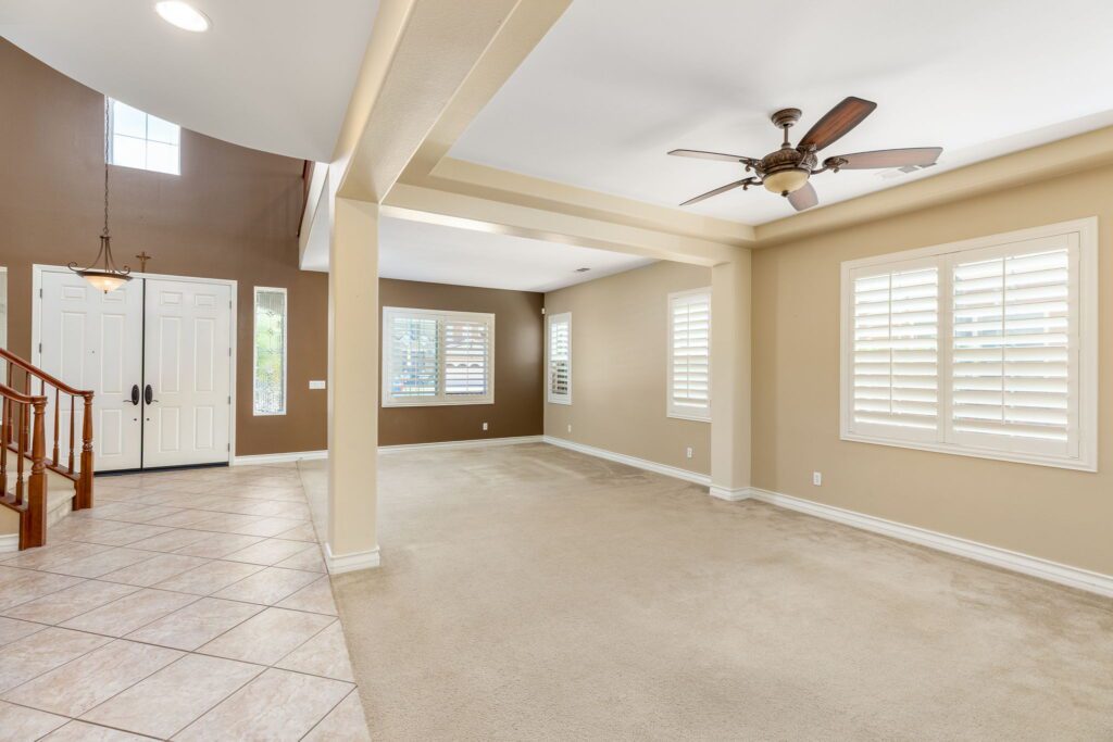 Spacious living area featuring light-colored walls, carpeted flooring, and plantation shutters. A ceiling fan and elegant staircase enhance the inviting atmosphere.