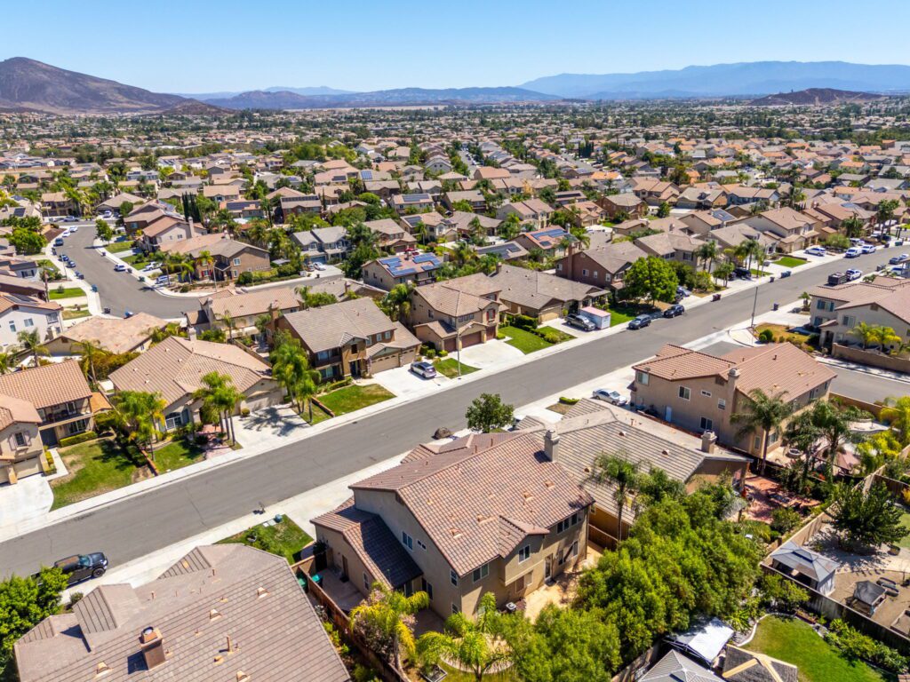 Aerial view of a suburban neighborhood featuring numerous homes with tile roofs, palm trees, and winding streets, set against a mountainous backdrop.