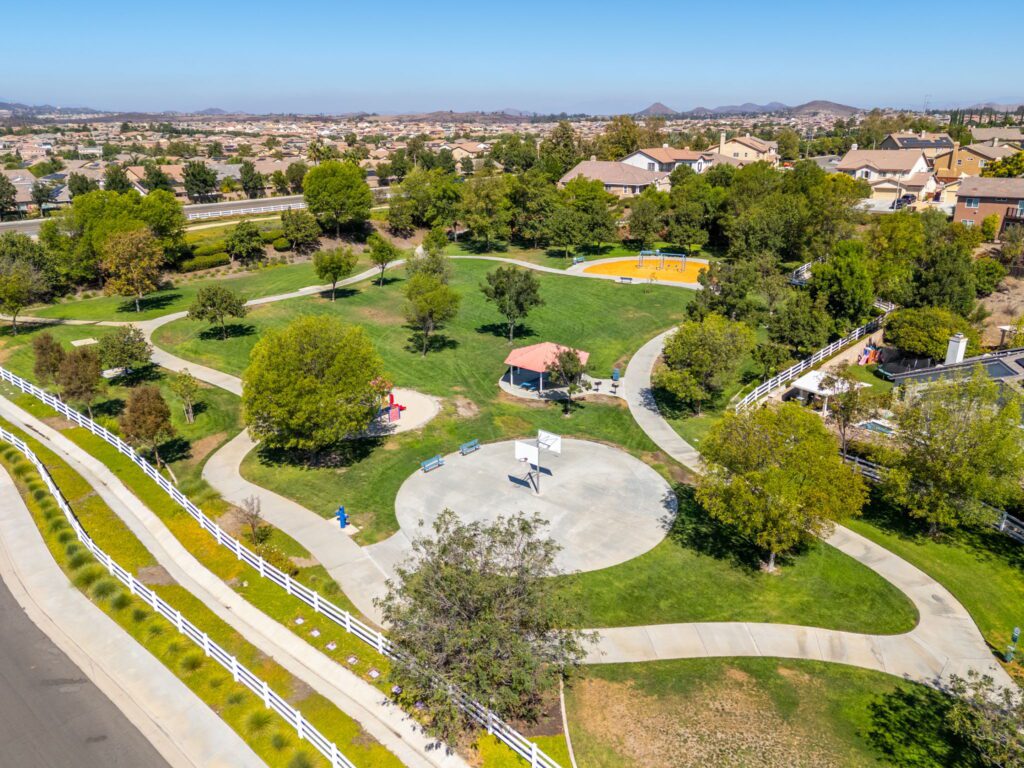 Aerial view of a community park featuring green lawns, walking paths, a playground, and a basketball court, surrounded by residential homes.