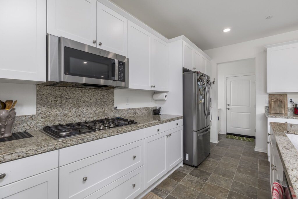 Bright, modern kitchen featuring white cabinets, granite countertops, stainless steel microwave, and a gas stove, with a refrigerator and entryway visible.