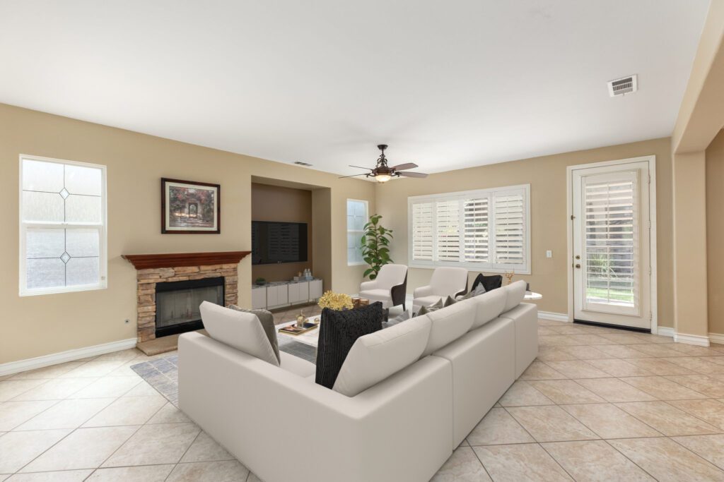 Bright living room featuring a white sectional sofa, a stone fireplace, and a flat-screen TV. Natural light streams through plantation shutters.