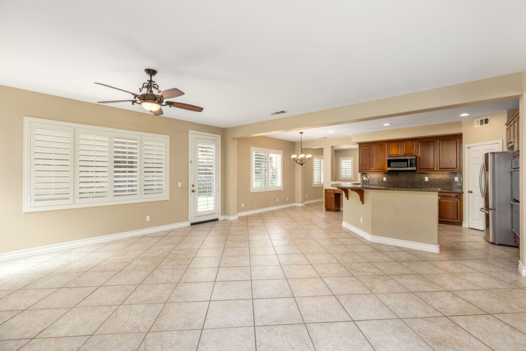Spacious open-concept living area featuring tiled flooring, a ceiling fan, and a view into the kitchen with dark wood cabinetry. Natural light floods through large windows.