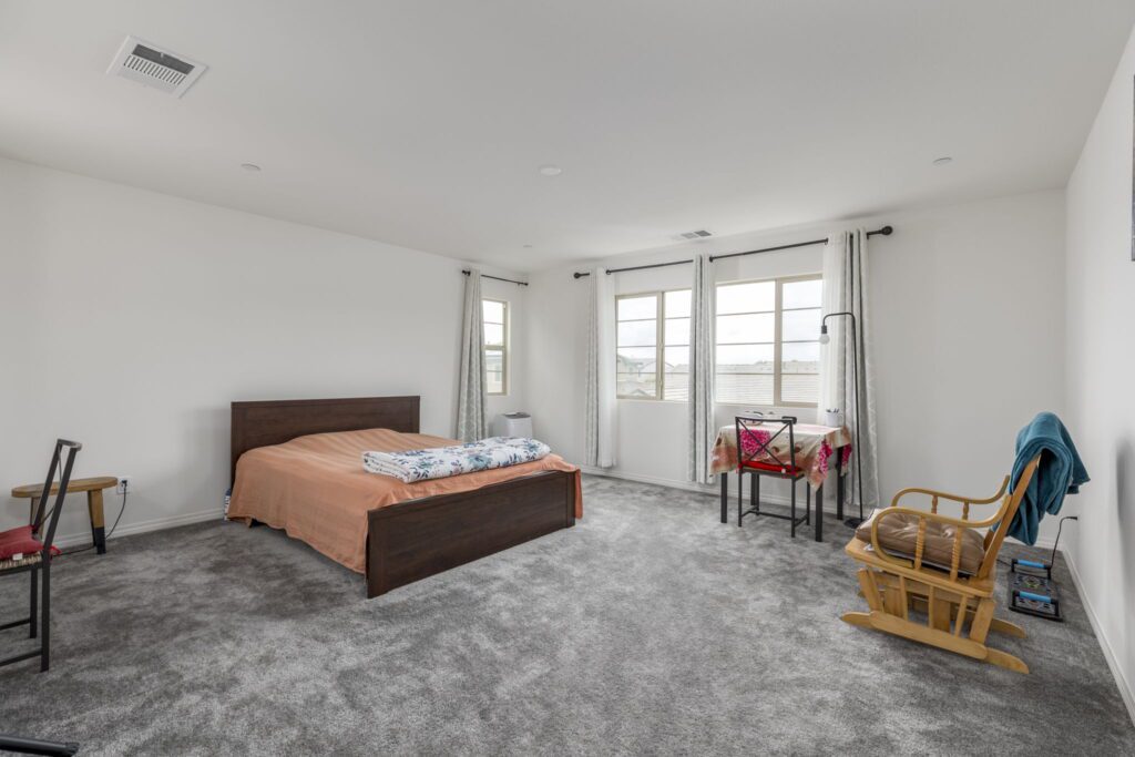 Spacious bedroom featuring a wooden bed with a light orange bedspread, large windows, a rocking chair, and a small table. Soft carpet enhances comfort.