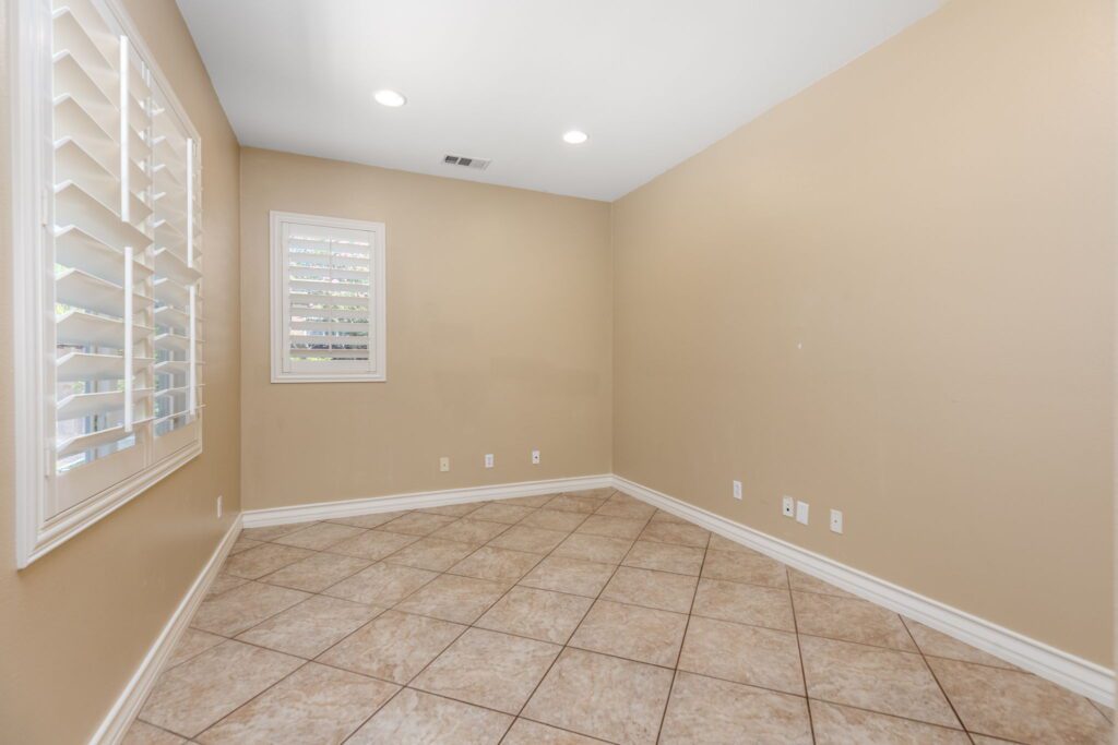 Empty room with beige walls, tiled floor, and window shutters. Natural light enters, highlighting the spacious layout. Ideal for interior design ideas.