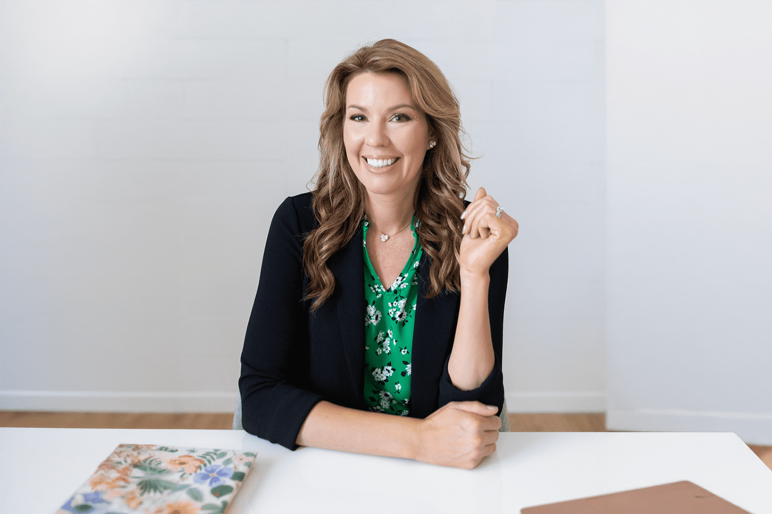 Woman in a green floral blouse and black blazer smiles while seated at a desk. The neutral background suggests a professional or creative environment.