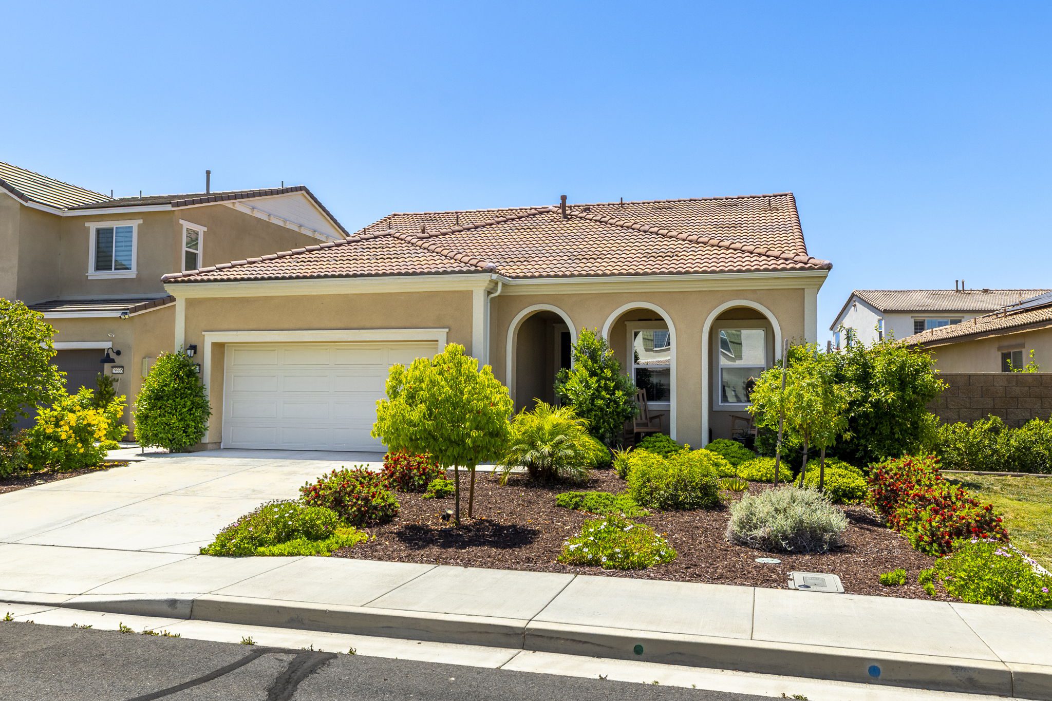 Single-story home with a tiled roof, featuring arched entryways, surrounded by lush landscaping and colorful flowers. Ideal for real estate listings.
