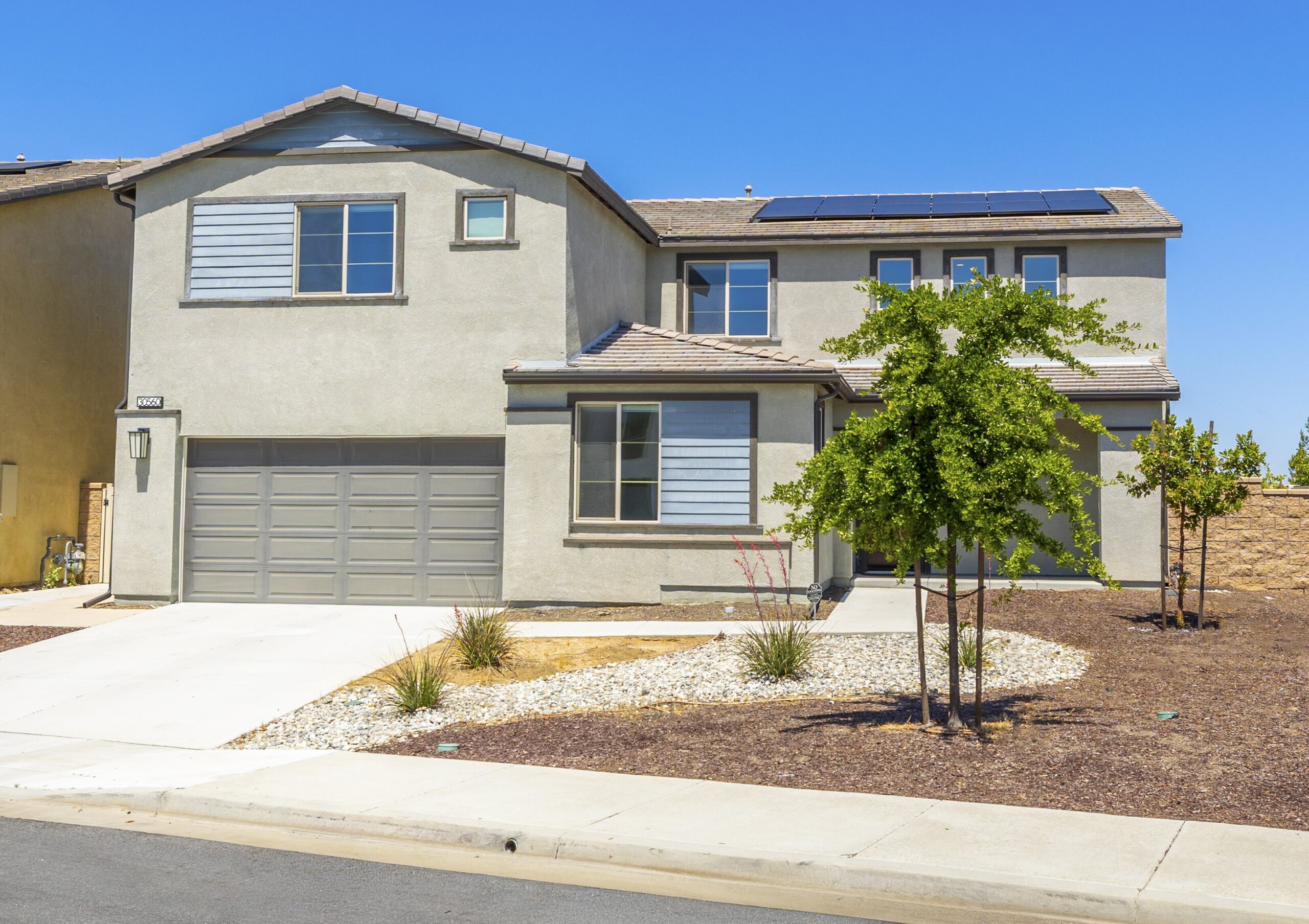 Two-story modern home with a gray exterior and solar panels, surrounded by landscaped yard and neighboring houses on a sunny day.