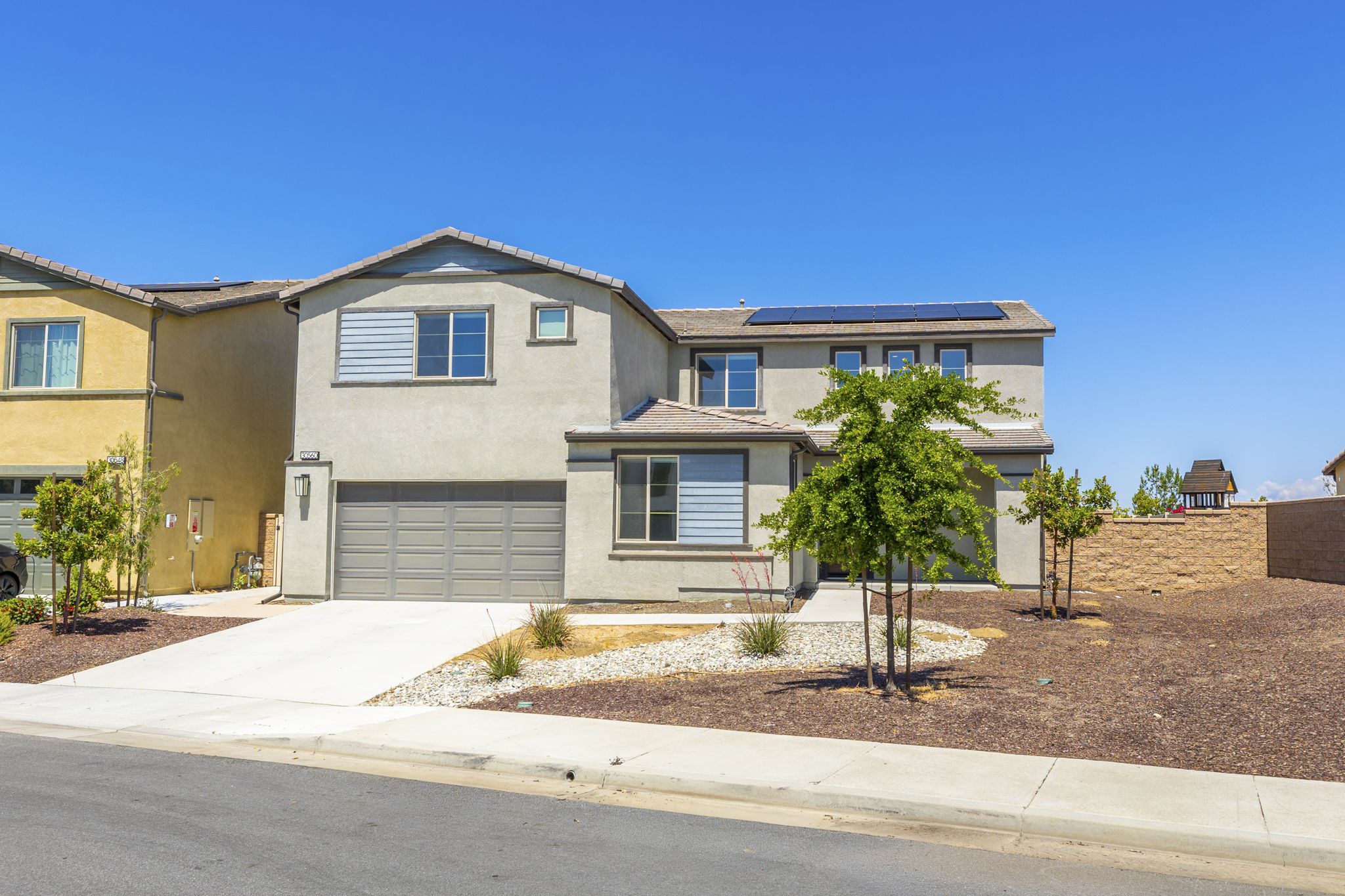 Two-story modern home with a gray exterior and solar panels, surrounded by landscaped yard and neighboring houses on a sunny day.