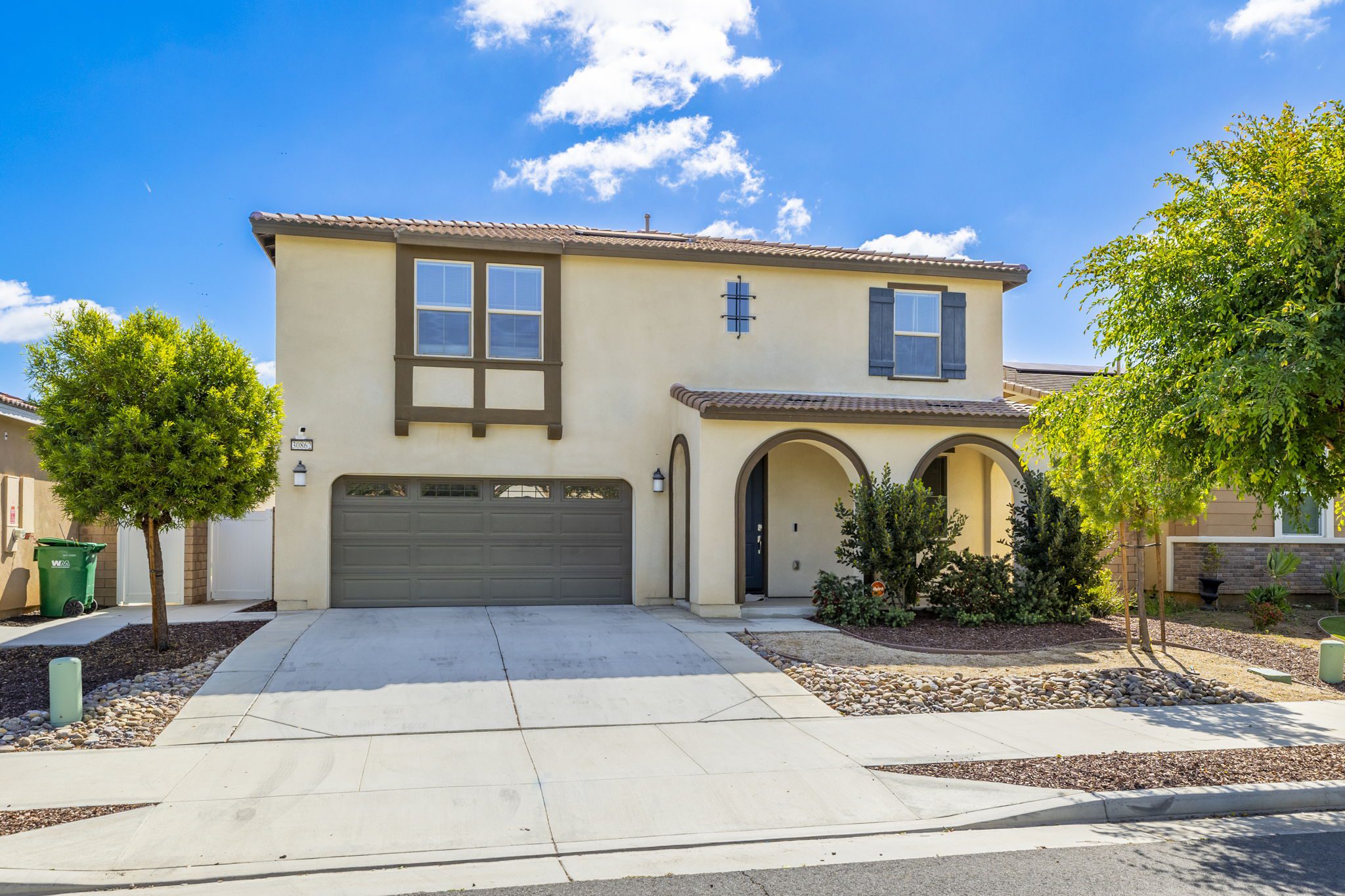 Two-story residential house with a beige exterior, dark shutters, and a landscaped front yard, showcasing a driveway and clear blue sky.