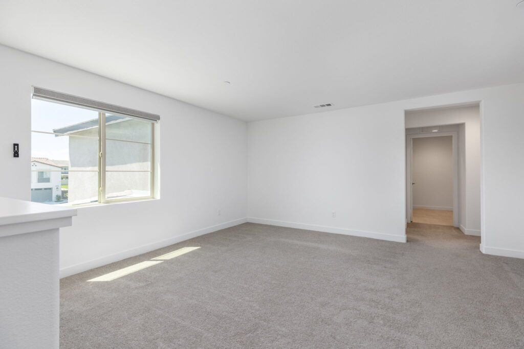 Bright, airy living room with light-colored carpet and a large window offering a view outside. A doorway leads to a hallway.