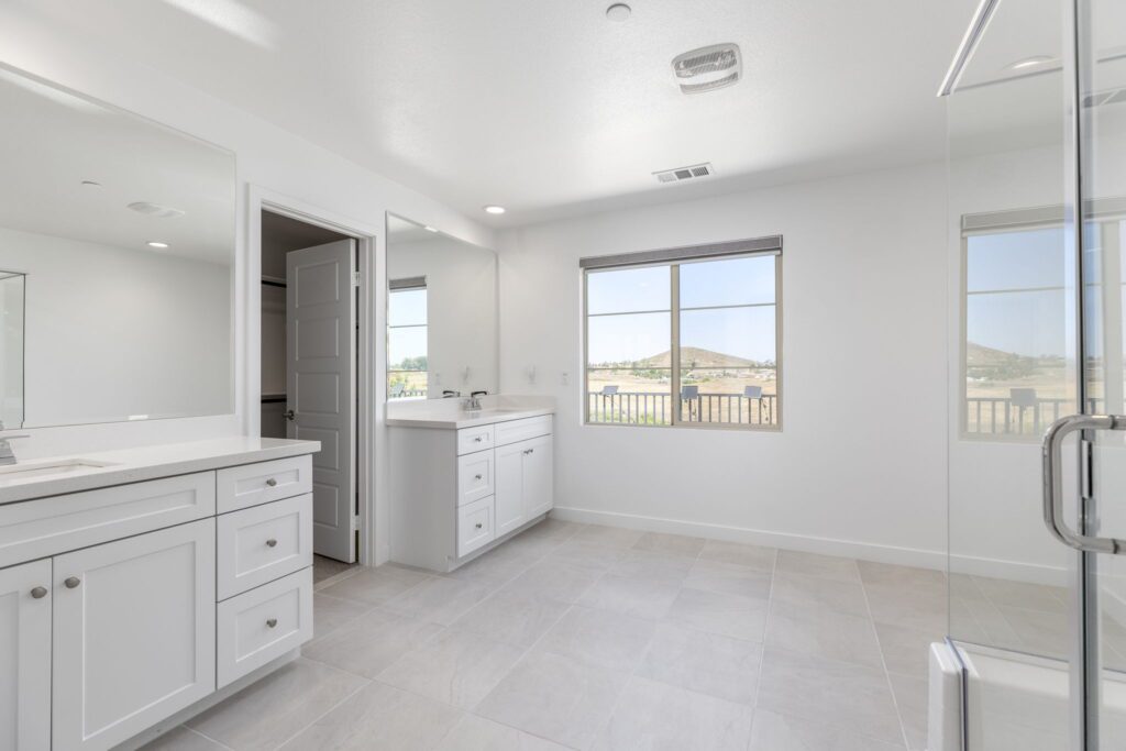 Spacious, modern bathroom featuring dual vanities, large mirrors, and a window with a scenic view. Sleek tile flooring completes the design.