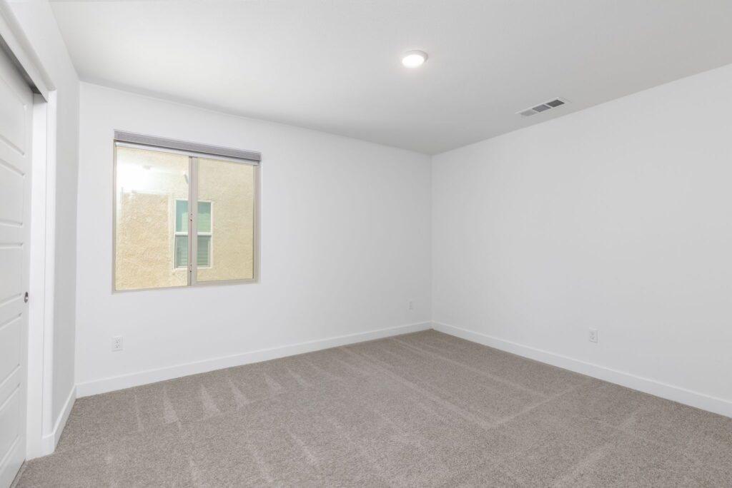 Spacious, empty room featuring light gray carpet, a large window showing a beige wall outside, and minimalistic white walls, ideal for customization.