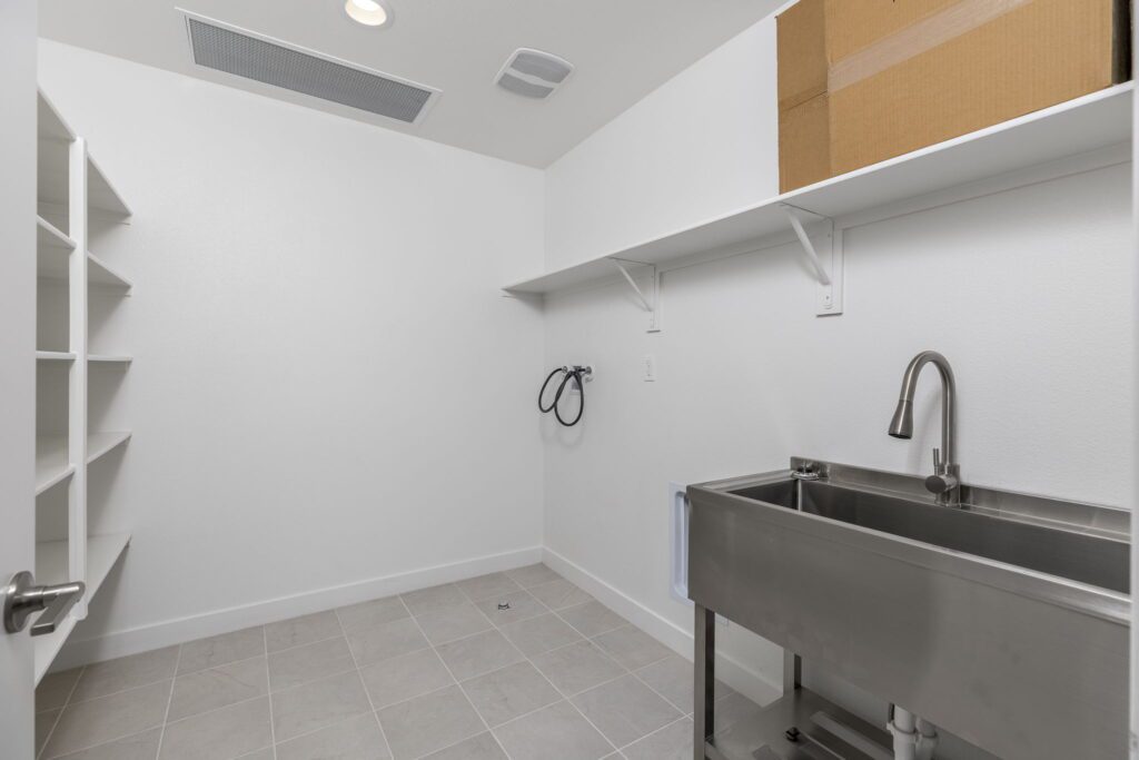 Laundry room featuring a stainless steel sink, wall shelves, and tile flooring, emphasizing spaciousness and utility for chores.