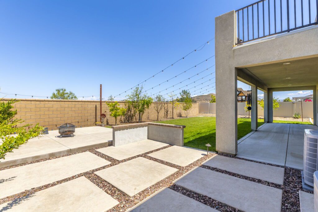 Backyard view featuring a stone patio, fire pit, and grassy area with decorative plants. String lights hang above, creating a cozy atmosphere.