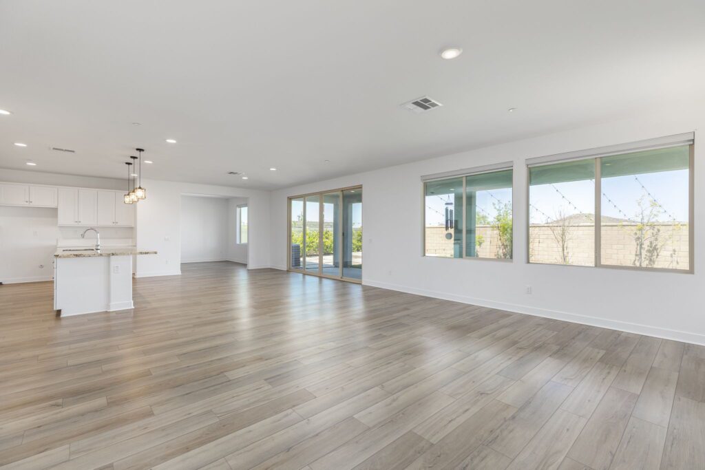 Spacious, sunlit living area with modern design, featuring large windows and wooden flooring, connecting to a kitchen with a white cabinetry.