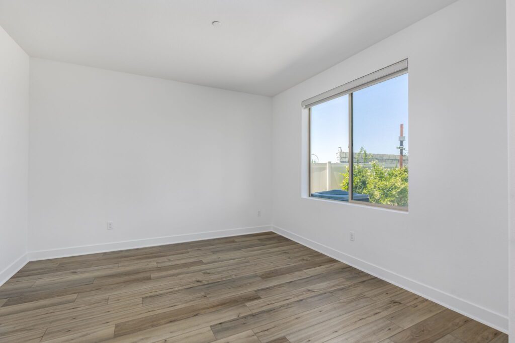 Bright, empty room featuring light wood flooring and large window with outside greenery visible. Ideal for showcasing apartment interiors or staging.