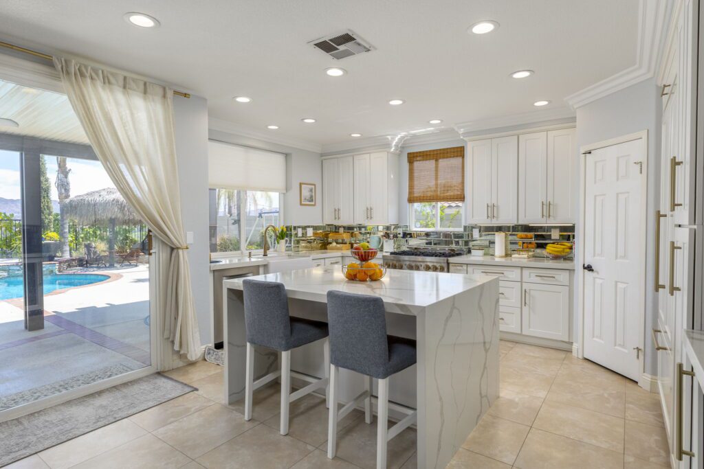Bright, modern kitchen featuring a large island with fresh fruit, white cabinetry, and a view of a poolside patio. Ideal for entertaining.