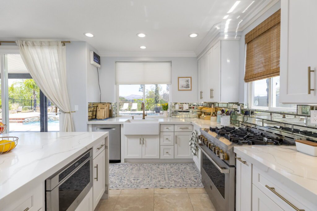 Bright, modern kitchen with white cabinetry, a large farmhouse sink, and a stainless-steel gas range. Sunlight streams in, revealing an outdoor pool area.
