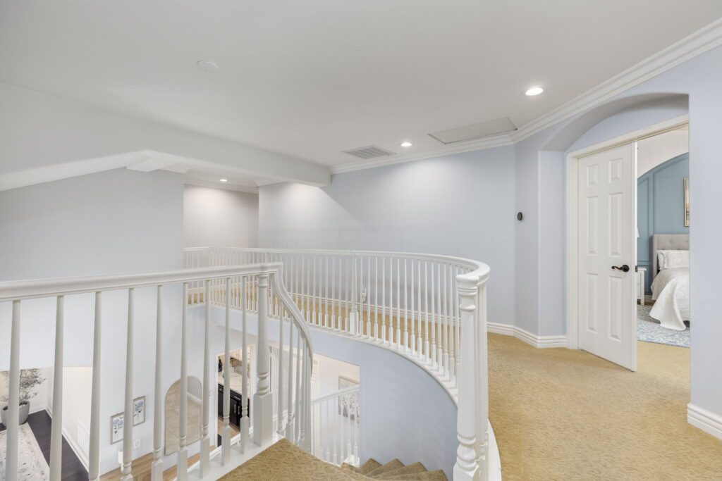 Bright, airy upstairs hallway featuring white railings and beige carpet. A doorway leads to a well-decorated bedroom, enhancing the home's inviting atmosphere.