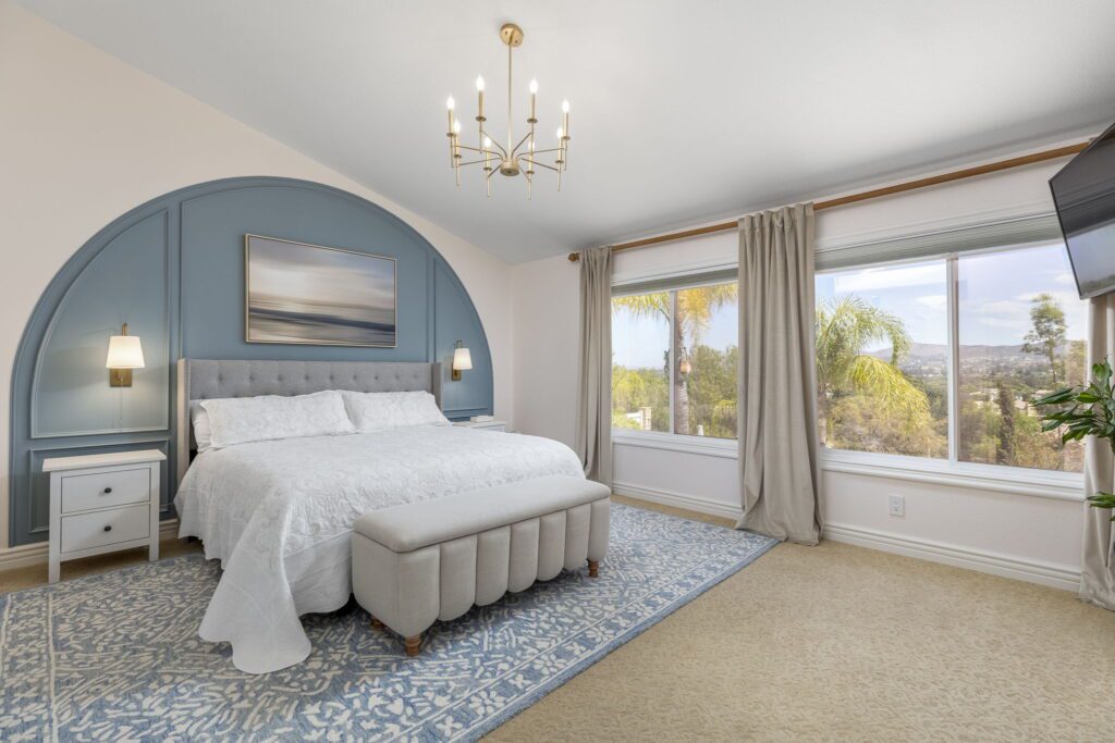 Bright and modern bedroom featuring a tufted gray bed, chic blue accent wall, and large windows with a scenic view. Ideal for showcasing interior design.