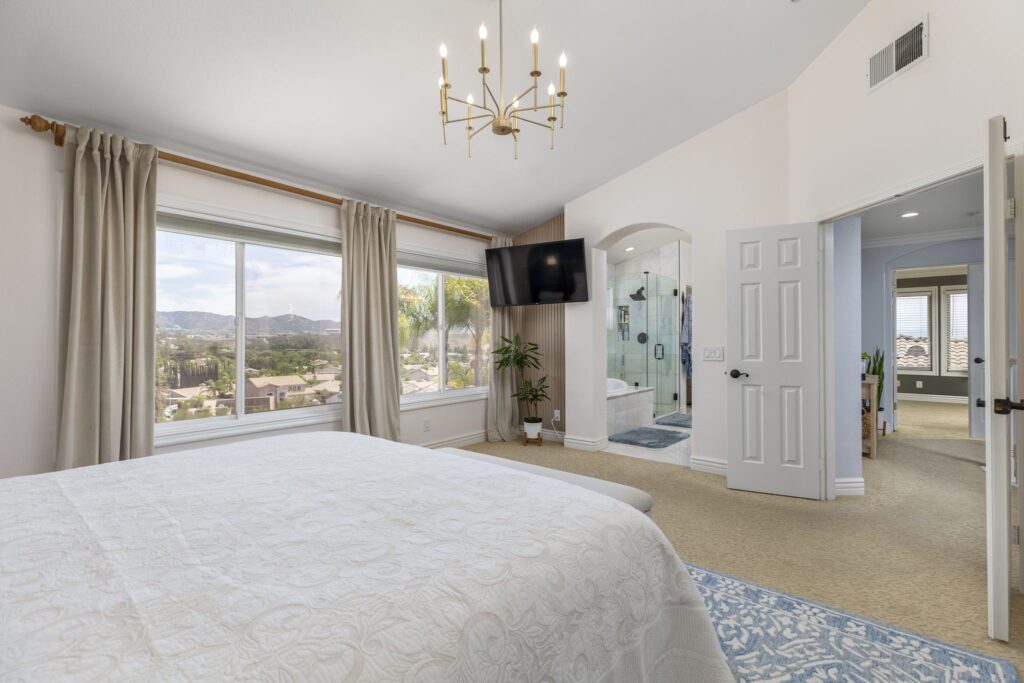Bright, modern bedroom featuring a large window with mountain views, a chandelier, and an ensuite bathroom visible through an open door.