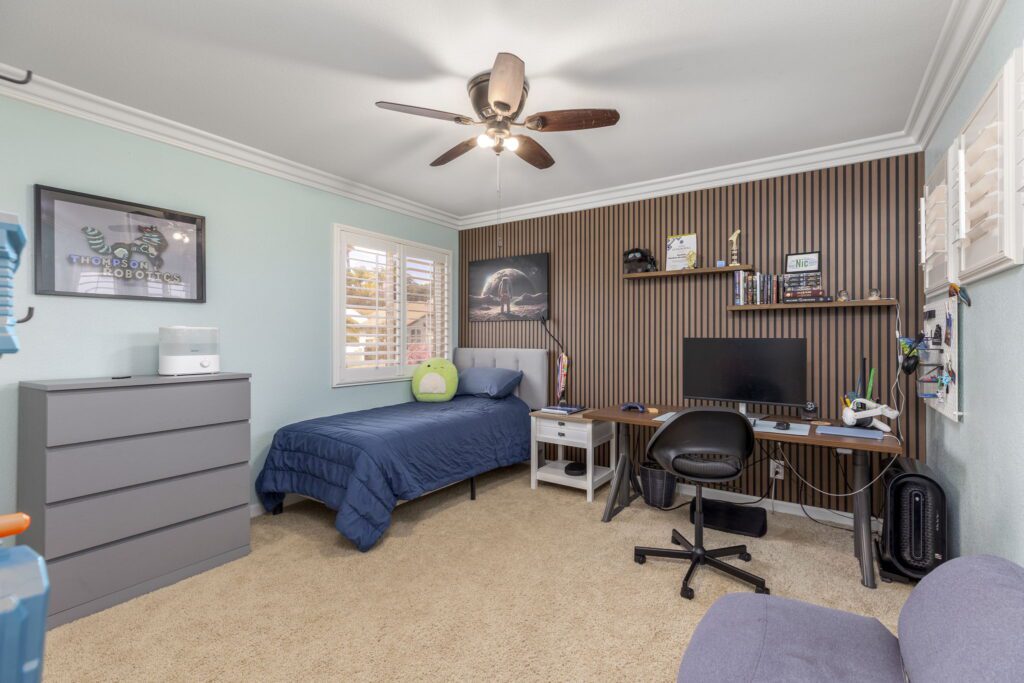 Modern bedroom featuring a blue quilted bed, a study desk with a computer, and a wall-mounted shelf displaying memorabilia, promoting functionality and style.