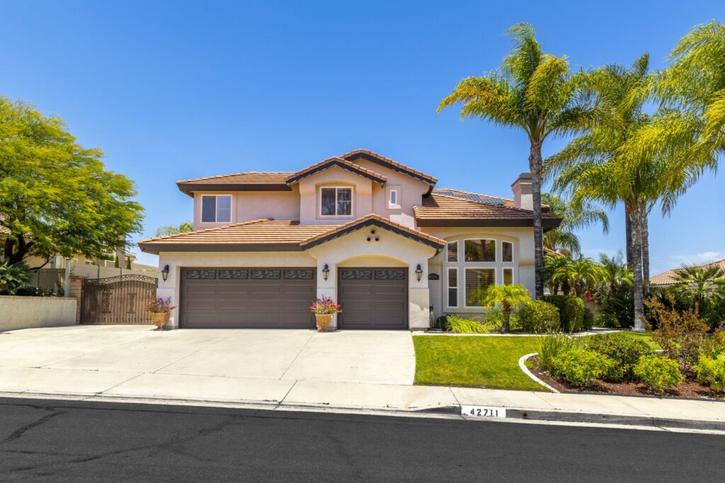 Two-story residential home with a tiled roof, surrounded by palm trees and greenery. Driveway leads to a triple garage, showcasing a sunny exterior.